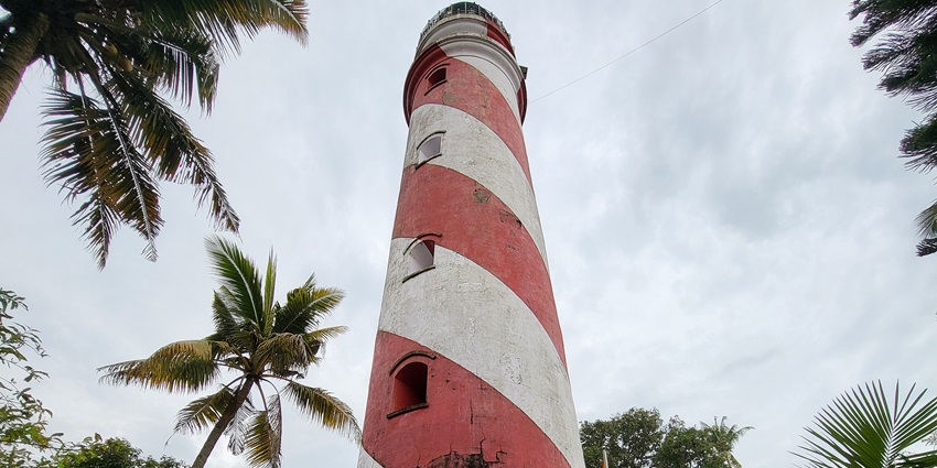 A picture of a light house at a sea shore in Kerala, surrounded with pine trees