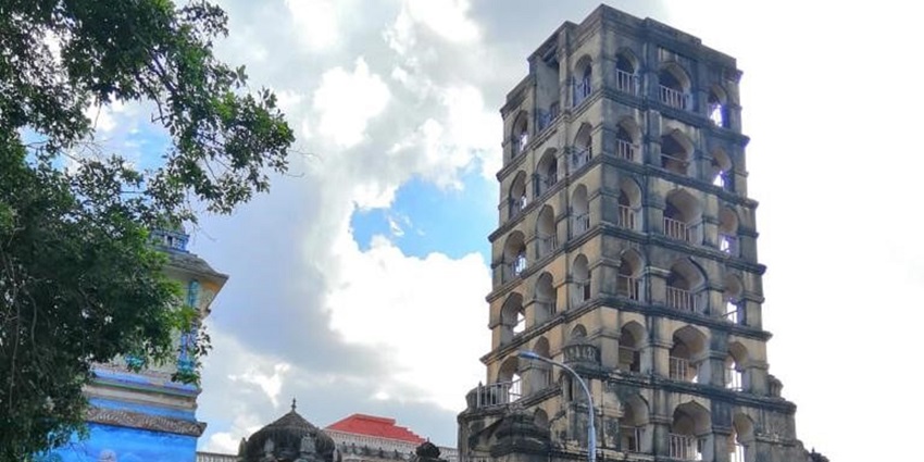 A distant picture of a bell tower in the middle of a street in Daman, surrounded by trees.