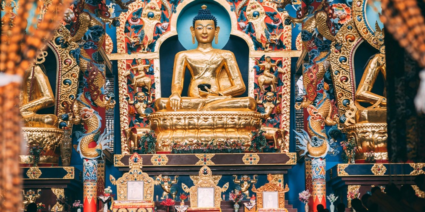 Image of the gold buddha statue in a gompa with beautiful decoration and devotees praying.