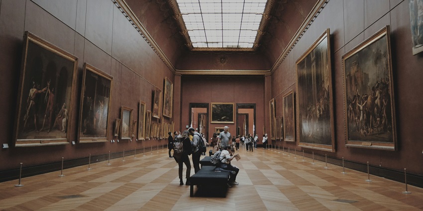 People walking through a gallery corridor, observing paintings displayed on the walls.