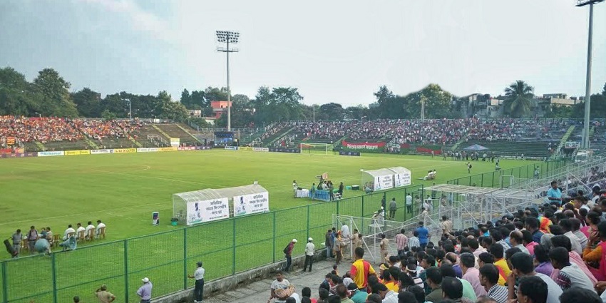 Children playing football on a grassy sports field with trees and buildings in the background