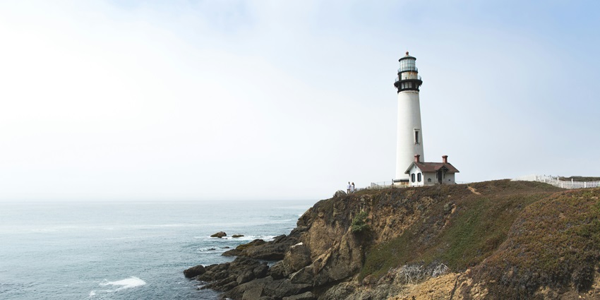 A distant view of a lighthouse on a small hill near a body of water during the daytime.
