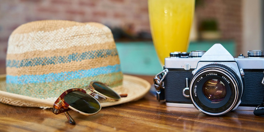 Grey and black DSLR camera rests beside a sun hat and sunglasses on a sunny surface.