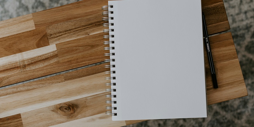An image of a white spiral notebook with blank pages & a pencil kept on a wooden table.