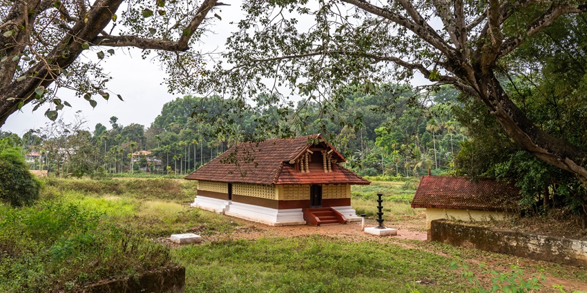A small Kerala-style Hindu temple with a red-tiled sloping roof, golden finials, and stone walls