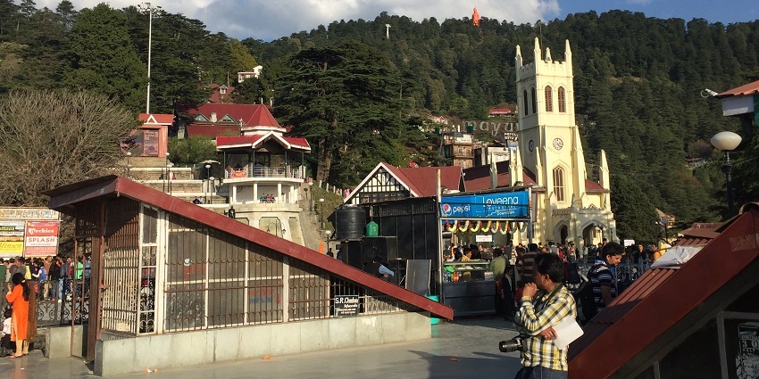 View of Jakhu temple from mall road in Shimla, surrounded by lush greenery under clear sky.