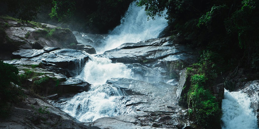 Waterfalls cascading over rugged rocks amidst a lush, dense forest backdrop.