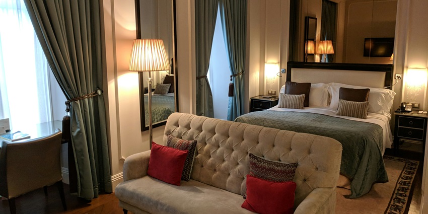 A picture of a hotel room with a couch placed in front of a bed with lit-up lamps