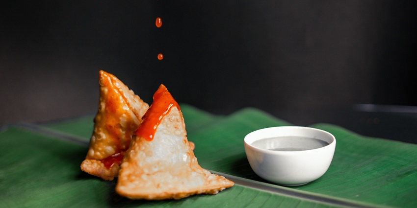 A mouth-watering view of a plate served with two samosas and a dip falling on top of it.
