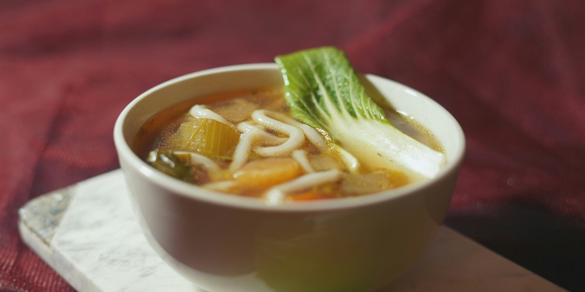 A delicious view of a bowl with noodle soup, vegetables, green celery and a spoon in it.