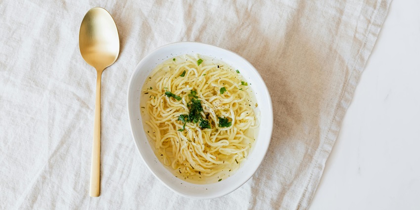 A tempting view of a bowl served with noodle soup kept on a white table beside a spoon.