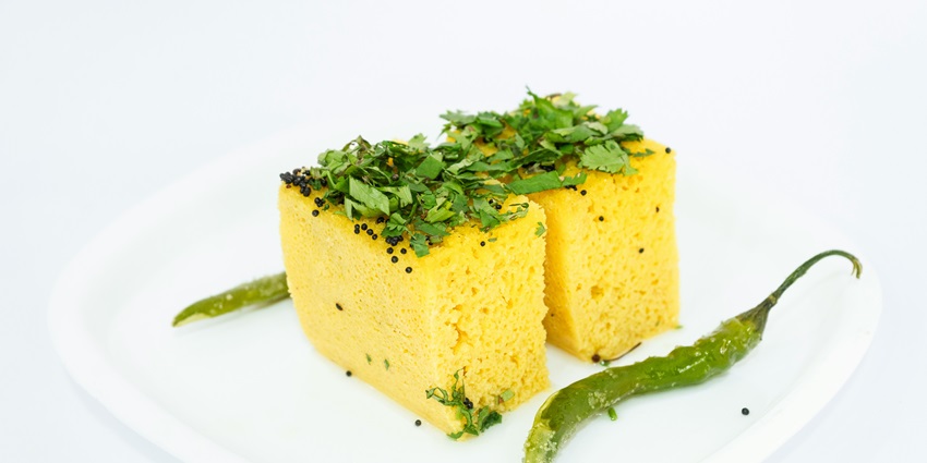 A mouthwatering view of a white plate served with yellow dhokla and garnished with greens.