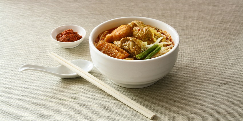 A mouth-watering view of a bowl of noodles with a spoon, chopsticks and a dip in the side.