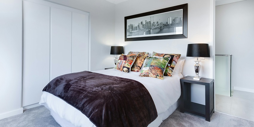 A landscape of a room with a bed lined with white bedsheets and colourful pillows on it.