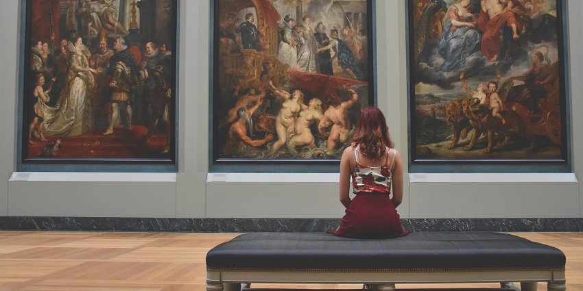 Woman sitting on an ottoman in front of three vibrant paintings in an art gallery.