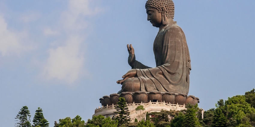 A picture of a tall Buddha statue in the sitting position around trees and greenery
