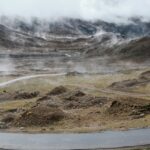 A view of a majestic mountain range with a narrow strip of road in between under clouds.