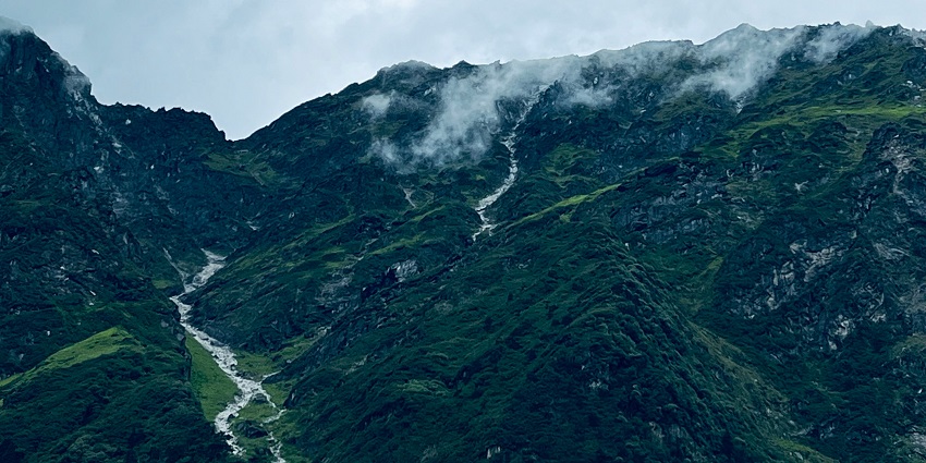 A view of a lush green mountain range with a stream of water flowing under clouds.