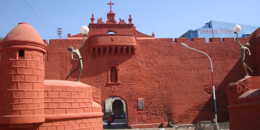 A frontal view of a majestic red building with a cross on its roof and lush surroundings