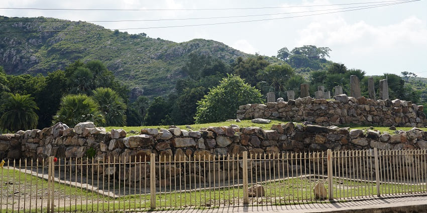 A picture of the ruins of a stupa with tall rocks standing at a grassy land