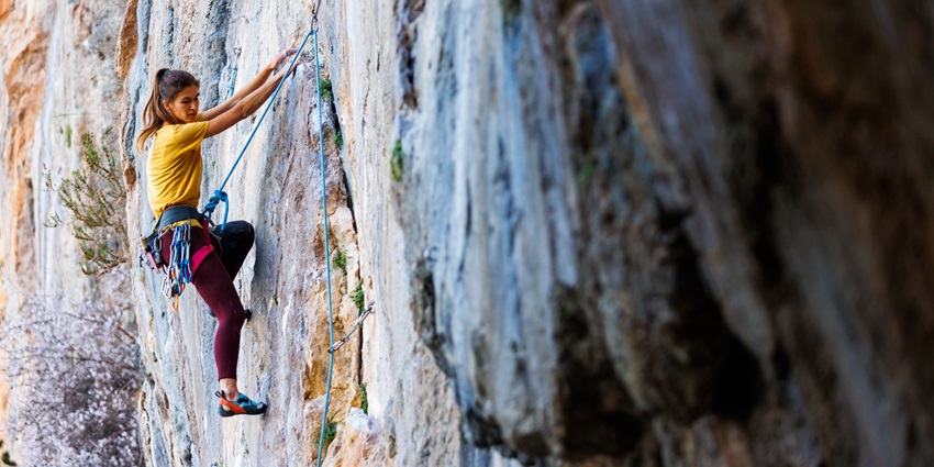 A person climbing a mountain with a steep rock face with a cloudy sky in the background.