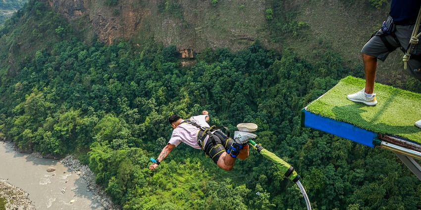 A man bungee jumping with safets gears on, with lush trees and river down in the cliff