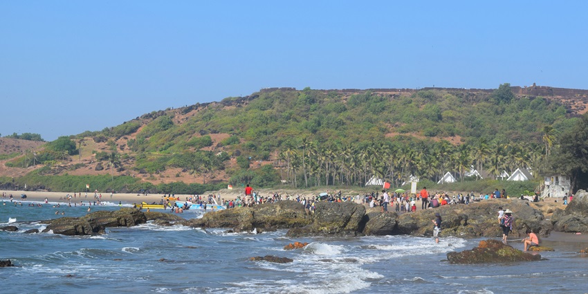 A distant view of the Chapora fort perched on top of a hill with tourists enjoying at a beach in front of it