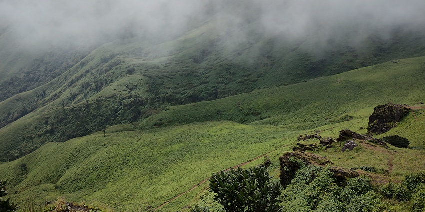 A picture of the vast green landscape taken from a hill with a cloud cover all over the place