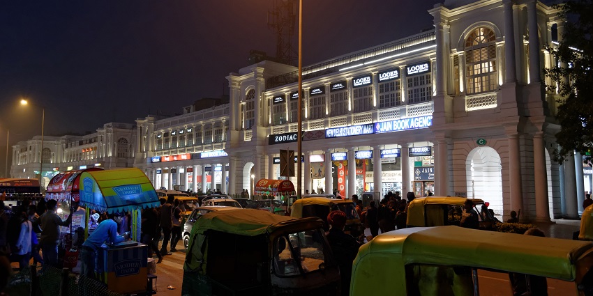 A picture of a bustling market in Delhi at night with vehicles parked around the roads