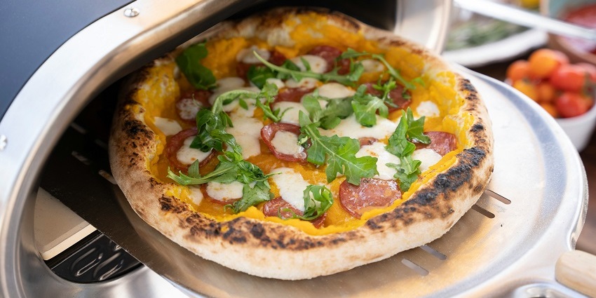 A mouth-watering view of a pizza being put into an oven for baking during the daytime.