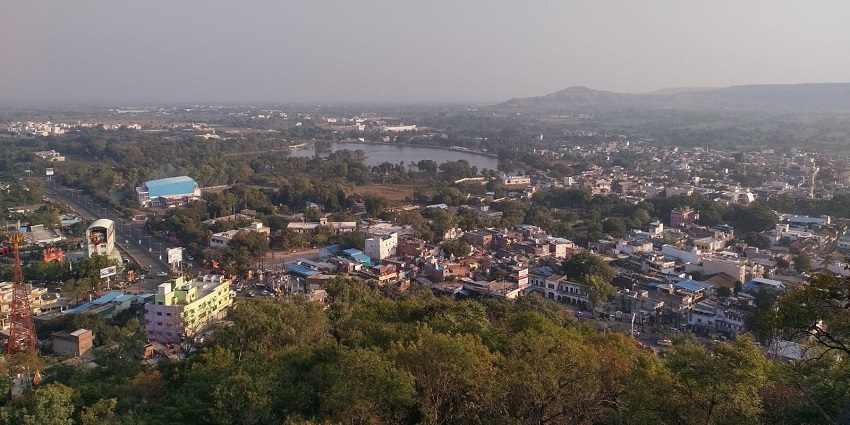 A panoramic view of Dewas with a river, greenery, and distant hills under a hazy sky