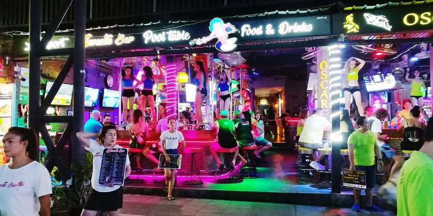 Host standing outside brightly neon-lit shops and bars, welcoming guests on Bangla Road