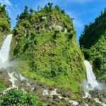 Image of twin waterfalls cascading through lush green rocks under clear blue sky