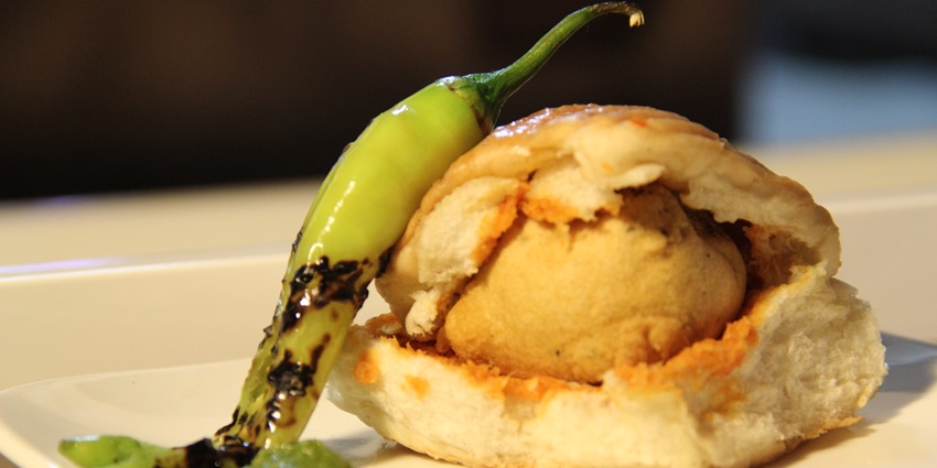 Image of a plate of vada pav served on a plate with a green chili on on the side and green chutney