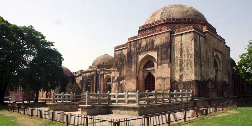 A picture of a historical tomb in Hauz Khas with a metal fence and lush greenery all around