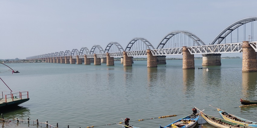 A picture of a long bridge passing through a river with a boat parked around the river shore
