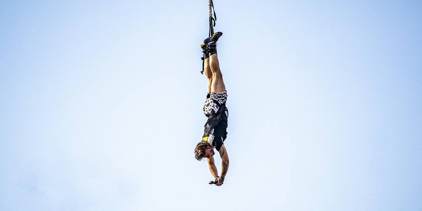 Man hanging on a rope high above a rocky landscape, with a blue sky in the background.
