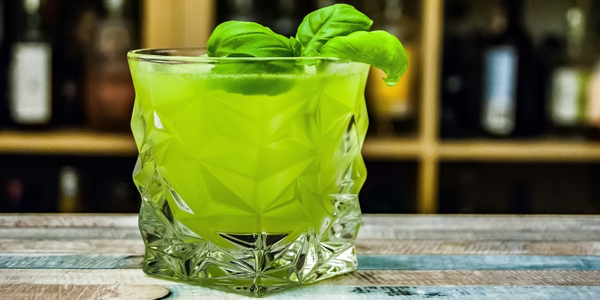 A close-up of a rock glass filled with vibrant green drink, set against a blurred background.