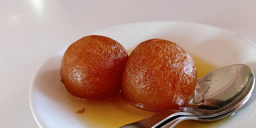 A delicious view of a plate served with two gulab jamuns with spoons on the side of it.