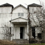 A glimpse of a desolate house standing on a barren land surrounded by dried trees.