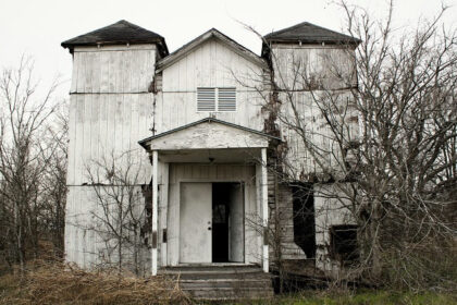 A glimpse of a desolate house standing on a barren land surrounded by dried trees.