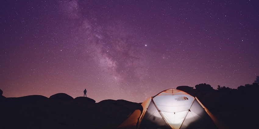 A man silhouetted near a tent, camping on a hilltop, with a scenic sunset in the background