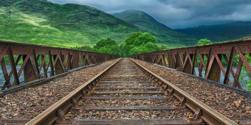 A brown train on a railway track, surrounded by greenery and a clear blue sky