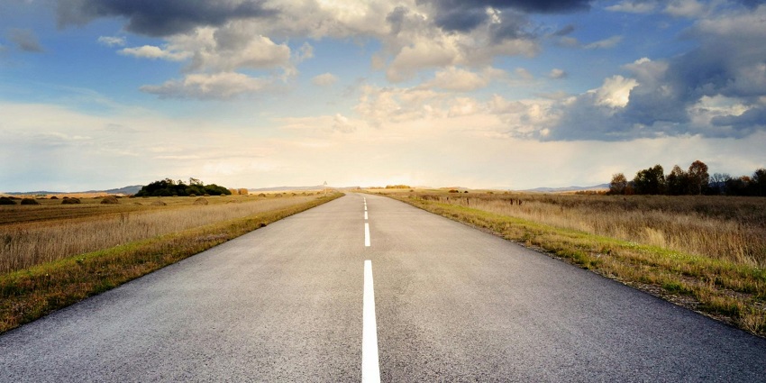 A road with fields on either side, under a cloudy sky during day time.