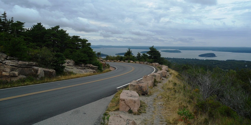 A mountainous road with cloudy skies overhead and greenery with some rocks on the side.