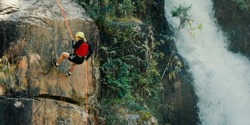 A woman climbing a rock near a waterfall, surrounded by lush greenery and flowing water.