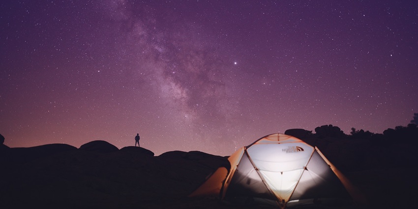 A tent pitched on a hillside with a man’s silhouette standing nearby, overlooking a scenic valley at sunset.
