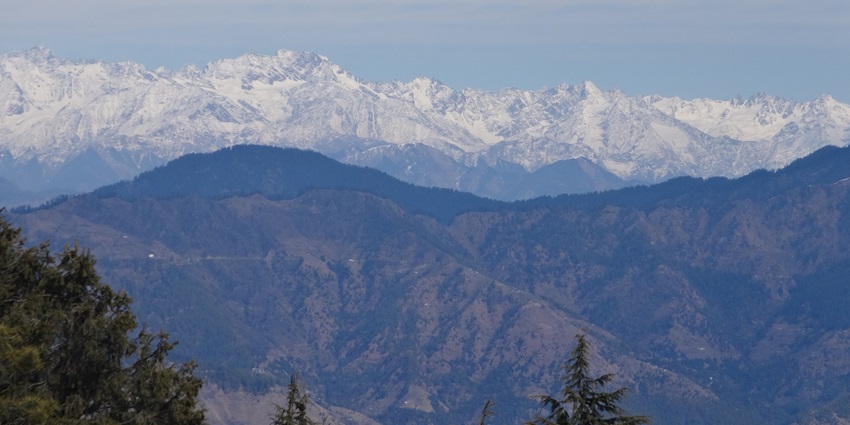 Scenic view of snow-covered Kufri hills surrounded by pine trees and mountains.