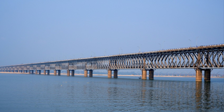 Long wooden bridge stretching over calm water with a backdrop of mountains and sky.