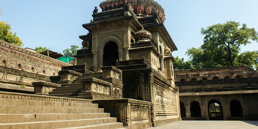 The Mahamrityunjaya Temple has intricate carvings and a prominent central spire, surrounded by lush greenery and a clear sky